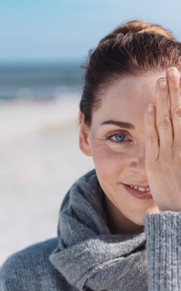 Eine junge lächelnde Frau am Strand hält sich ein Auge zu.