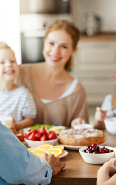 Eine junge Familie frühstückt zusammen am Esstisch. Mutter, Vater und drei Kinder lächeln in die Kamera.