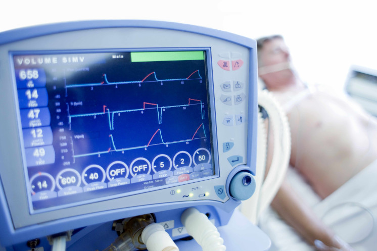 Bildschirm eines Überwachungsmonitors, im Hintergrund liegt ein Patient im Krankenhausbett